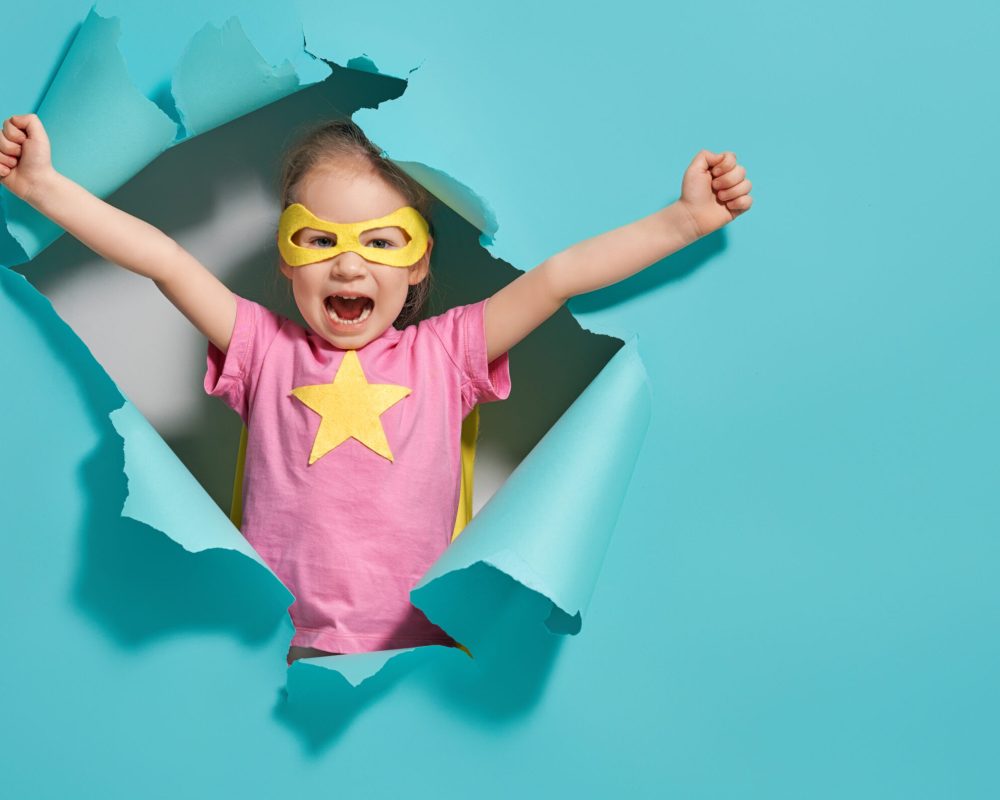 Little child playing superhero. Kid on the background of bright blue wall. Girl power concept. Yellow, pink and  turquoise colors.